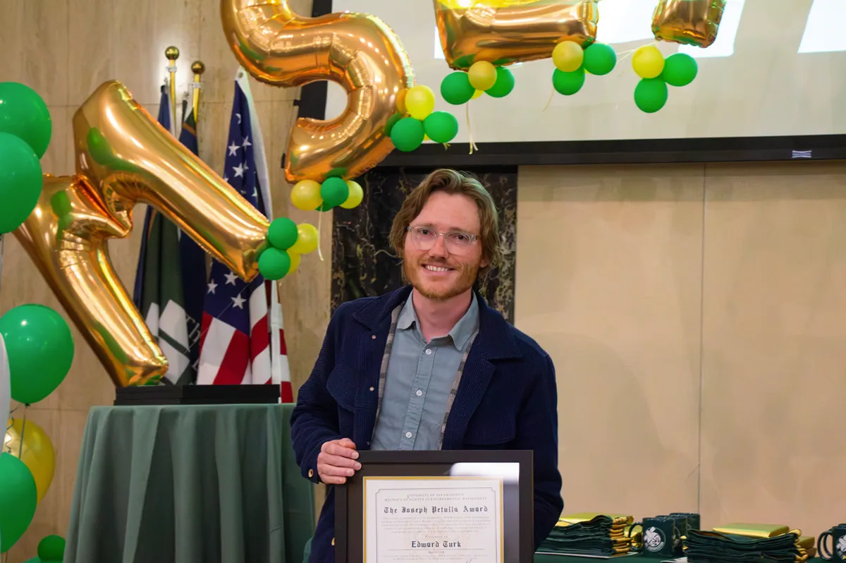 Edward Turk stands smiling behind a table, holding a framed certificate that reads The Joseph Petulla Award, Edward Turk. Large gold letter balloons and clusters of green and yellow balloons fill the background, with flags to one side and stacks of folded green graduation stoles on the table beside him.