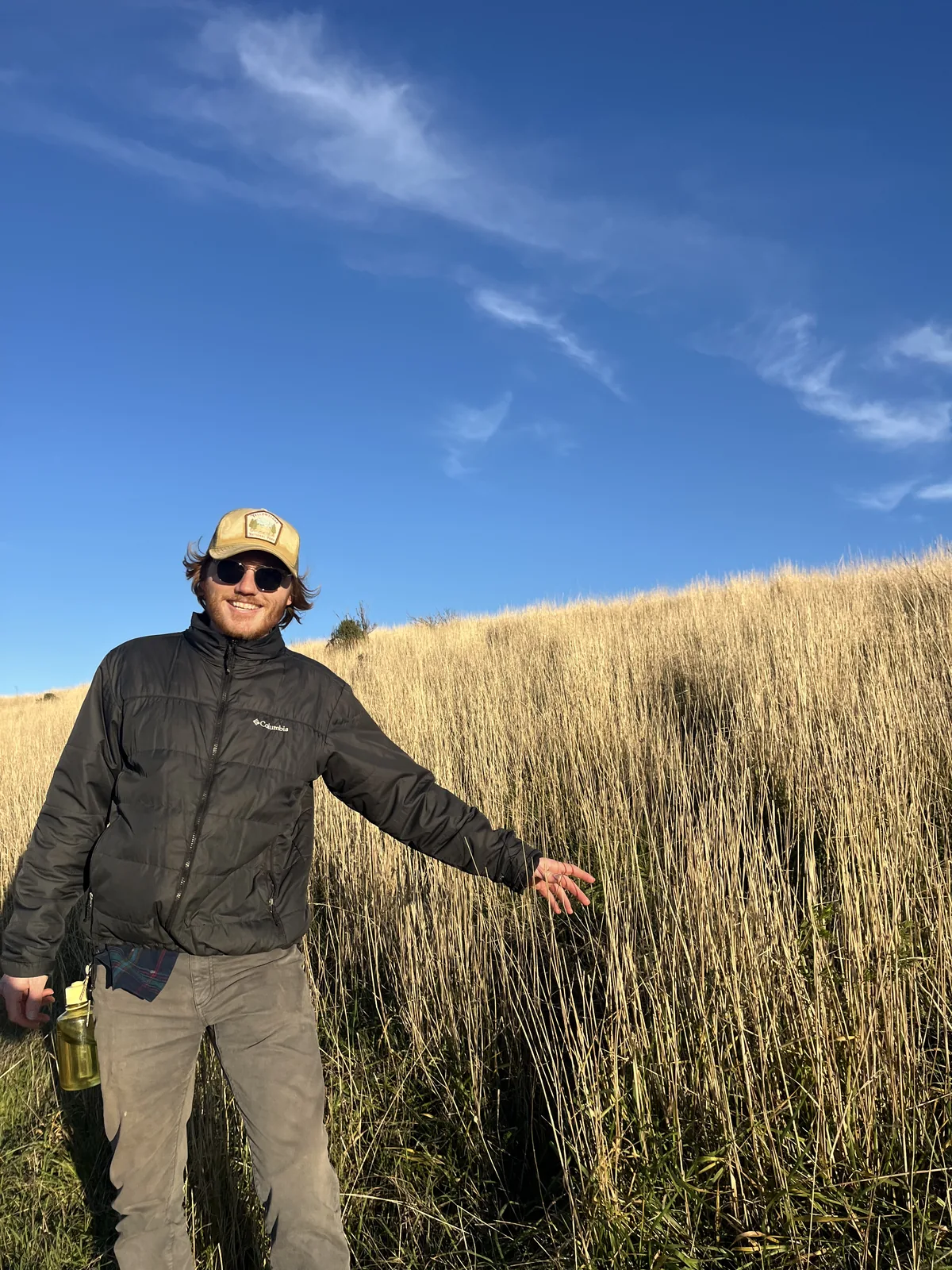 Edward Turk standing in a field of tall golden grass under a clear blue sky, wearing a cap and sunglasses and resting one hand on the grass.