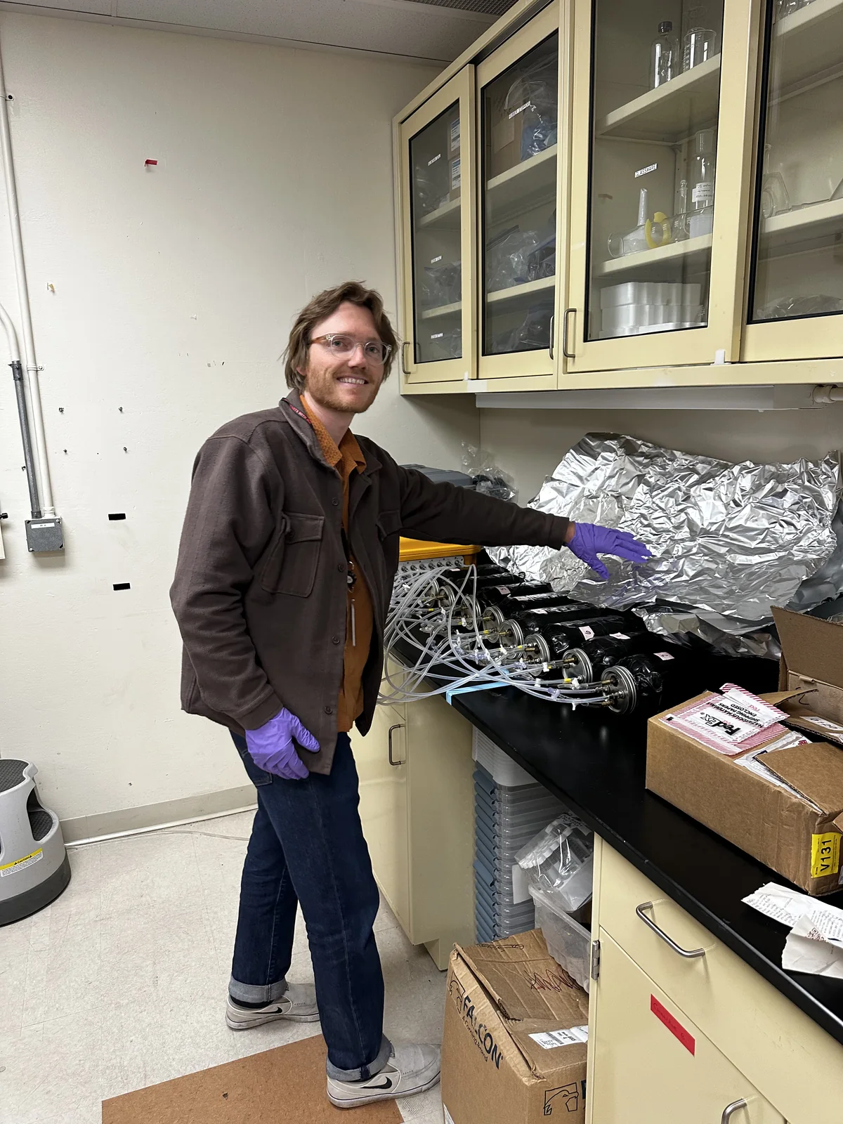 Edward Turk standing beside the incubation rig in the lab: the grey LI-7810 analyzer sits on top of the yellow LI-8250 multiplexer, with the row of foil-wrapped jars and gas lines on the bench.