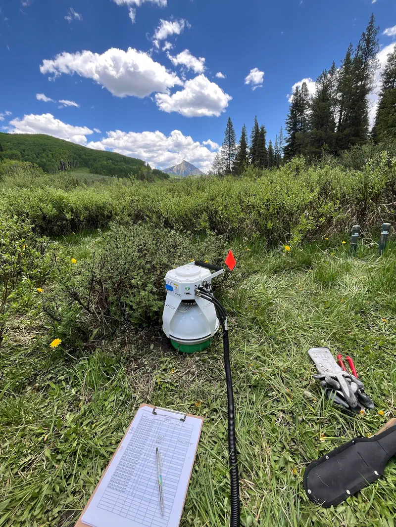 A white flux chamber sitting on grass at the edge of a willow thicket, with a ribbed hose, a marker flag, a clipboard holding a data sheet and a pair of work gloves beside it. Conifers and a pointed peak stand in the background under a blue sky.